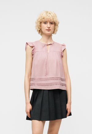 Žena s kovrčavom plavom kosom u ružičastoj bluzici bez rukava s naborima i crnoj plisiranoj suknji, stoji ispred jednobojne pozadine.