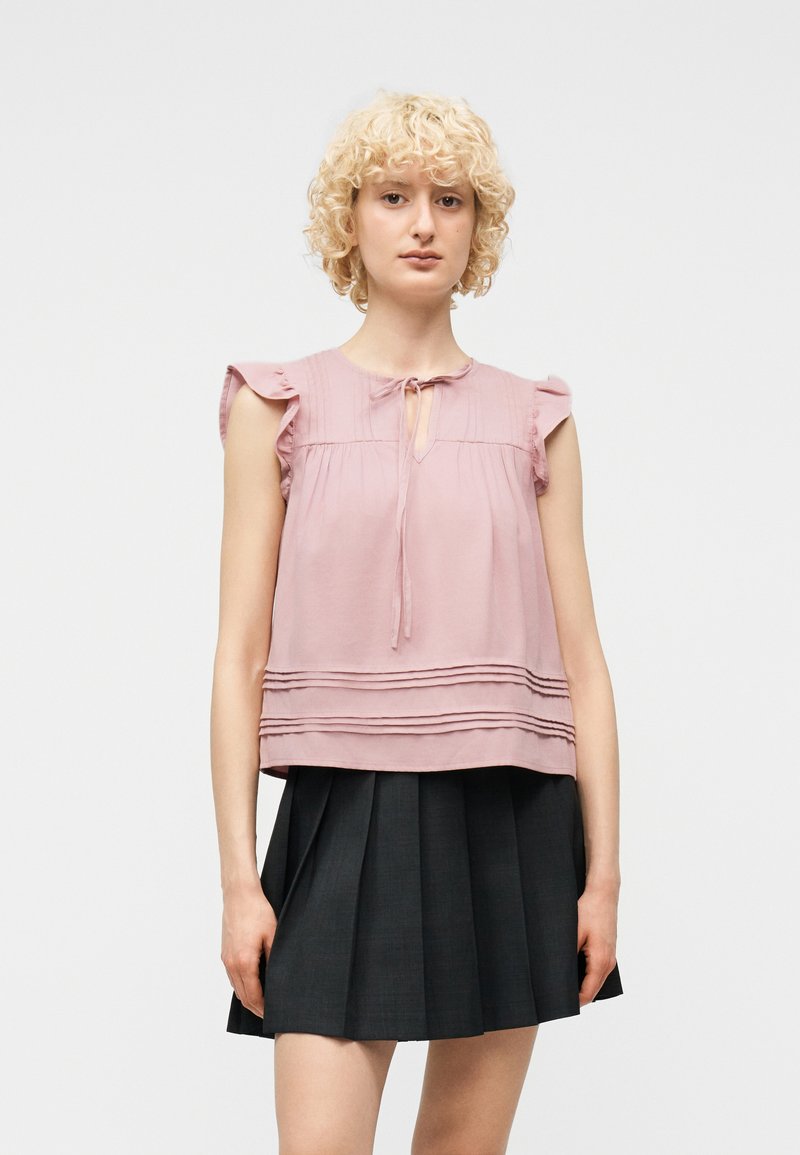 Γυναίκα με ξανθά σγουρά μαλλιά που φοράει ροζ αμάνικο τοπ με βολάν και μαύρη πλισέ φούστα, στέκεται μπροστά σε απλό φόντο.