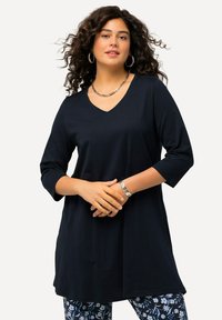 Tummansininen, v-kaula-aukkoinen tunika, jossa on kolmen neljänneksen hihat, valmistettu sileästä kankaasta. Mallilla on yllään kuviolliset housut ja hopeisia asusteita.