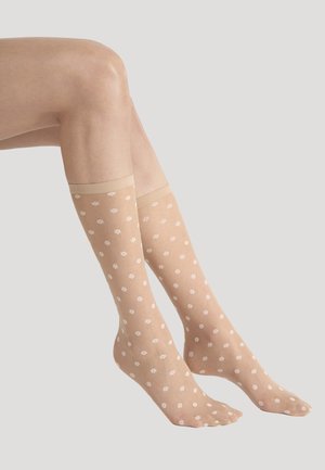 Ben med genomskinliga beige knähöga strumpor med små vita blomster mönster mot en enkel ljus bakgrund.