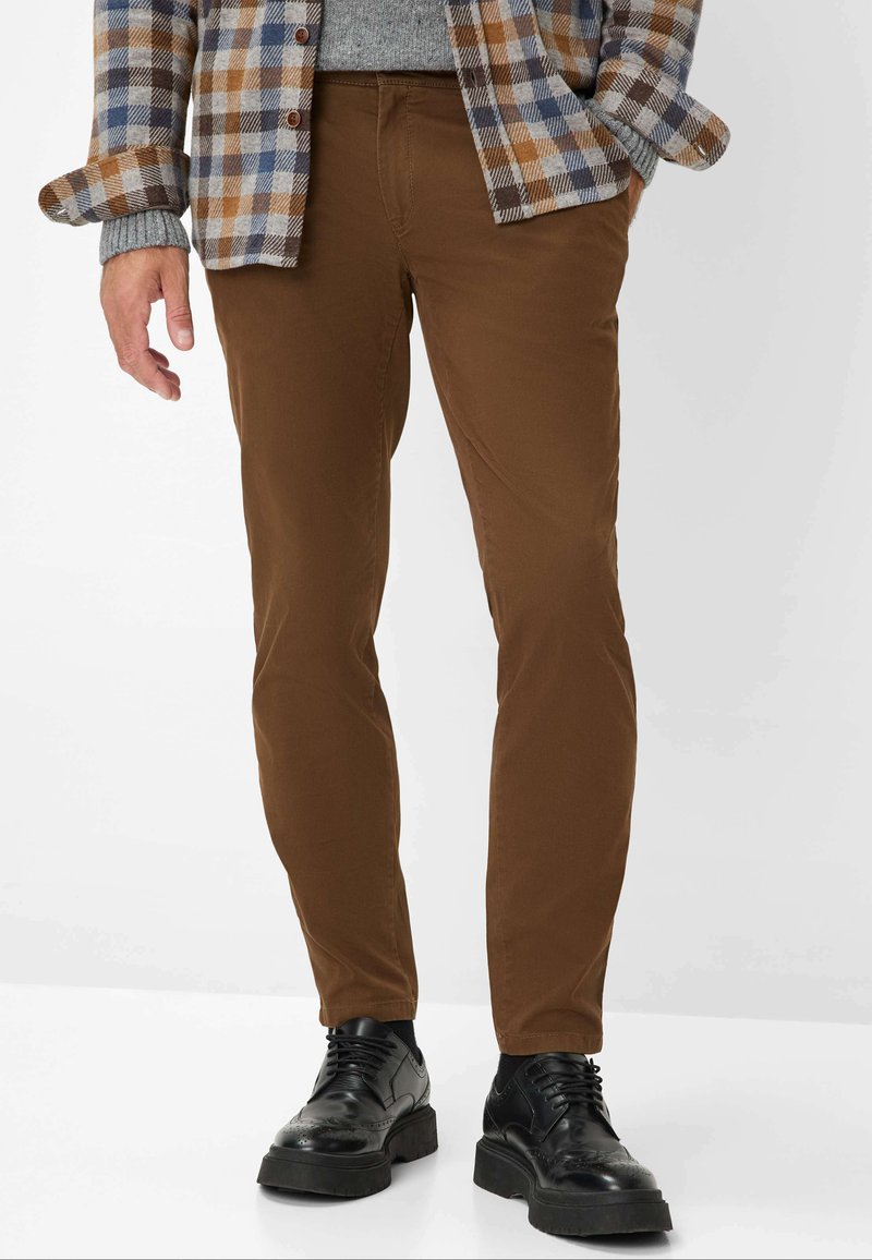 Pantaloni marroni slim-fit in un tessuto liscio. Abbinati a scarpe nere stringate. Camicia a quadri visibile sopra.