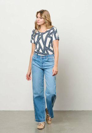 Vrouw die binnenshuis loopt, gekleed in een korte mouws top met geometrische print, lichtblauwe spijkerbroek met zijstrepen en beige veterschoenen.