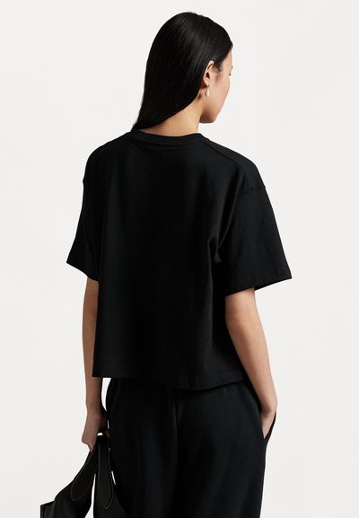 Tricou scurt negru, realizat dintr-un material moale, având mâneci scurte și o tivă dreaptă, asortat cu pantaloni negri și o geantă.