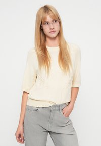 Jeune femme aux longs cheveux blonds portant des lunettes transparentes, un pull à manches courtes crème avec une broderie florale, et un jean gris clair avec une main dans la poche.