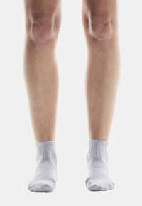 Calzini bianchi alla caviglia con polsini a coste, indossati su gambe nude, parzialmente visibili dalle ginocchia in giù, su uno sfondo bianco uniforme.