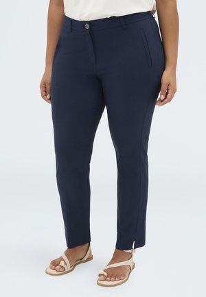 Kvinde iført navyblå tætsiddende bukser og beige sandaler, stående mod en ensartet lys baggrund.