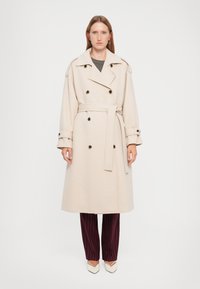 Beige Doppelreiher-Trenchcoat mit großen schwarzen Knöpfen, einem Gürtel in der Taille und breiten Revers; getragen über dunklen gestreiften Hosen.