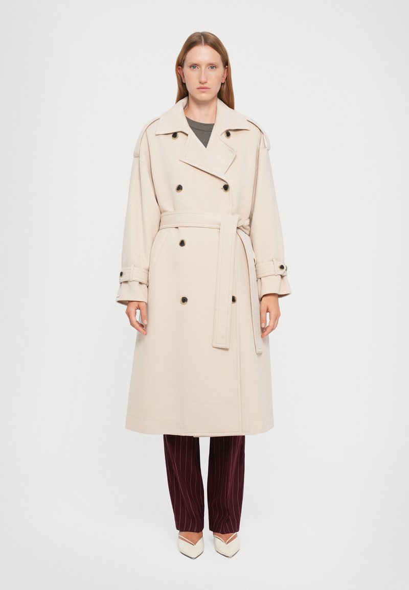 Beige Doppelreiher-Trenchcoat mit großen schwarzen Knöpfen, einem Gürtel in der Taille und breiten Revers; getragen über dunklen gestreiften Hosen.