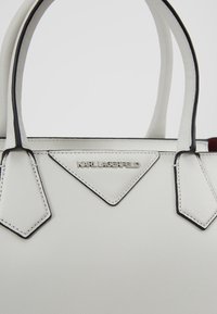Sac à main blanc avec une texture lisse, présentant des accents noirs et un élément de design triangulaire. Plaque de logo affichée de manière proéminente.