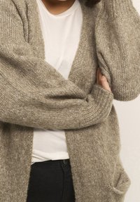 Béžový pletený cardigan s volnějším střihem, se širokými rukávy, hlubokým výstřihem do V a dvěma předními kapsami, navrstvený přes bílou košili.
