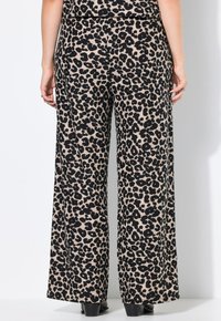 Wijduitlopende broek in een beige stof met een zwart luipaardprint patroon. Soepele textuur met een hoge taille en uitlopende snit. Zwarte schoenen.