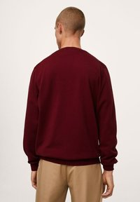 Sweatshirt bordeaux, texture tricotée, col rond, poignets et ourlet côtelés, coupe décontractée. Porté avec un pantalon beige, vu de dos.