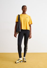Sárga crop top, fekete leggings mintás oldalsávval, és sárga/fekete sportcipő. A modell egy fekete bevásárlótáskát tart.