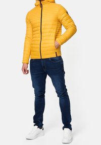 Gul polstret jakke med vandret syning, sort lynlås lukning og hætte, sammen med mørkeblå jeans og hvide sneakers.