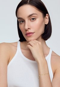 Złota bransoletka z małymi wiszącymi koralikami, noszona na nadgarstku. Modelka ma krótkie, ciemne włosy i ma na sobie białą bezrękawnik.