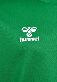 Tissu vert à surface texturée avec un logo "hummel" blanc et un motif d'abeille abstrait en dessous du texte.