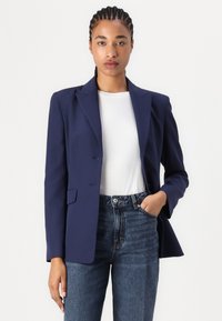 Marineblau blazer mit Revers, zwei Knöpfen und zwei Fronttaschen, kombiniert mit einem weißen Hemd und hochtaillierten Jeans.