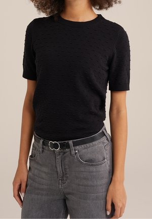 Kvinde iført en sort tekstureret kortærmet top, der er stukket ned i højtailede grå jeans med et sort bælte med dobbeltringsspænde.