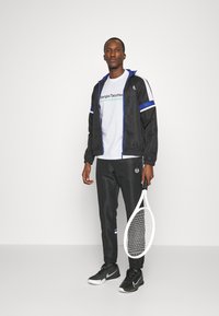Melna sporta treniņtērps ar ziliem un baltiem akcentiem, uzvilkts virs balta T-krekla ar "Sergio Tacchini" logo. Tur rokā tenisa rakete.