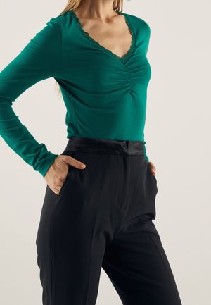 Kvinde iført langærmet teal top med rynket halsudskæring og blonder trim, stoppet ned i højtaljede sorte bukser med hænderne i lommerne.