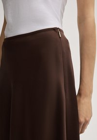 Jupe A-line marron en tissu lisse, dotée d'une fermeture éclair discrète sur le côté et d'une silhouette épurée. Associée à un t-shirt blanc.