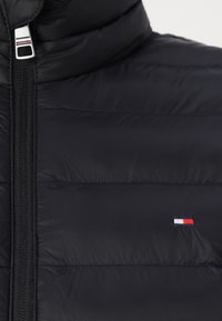 Black puffer jacket with a vertical zipper, featuring quilted texture and a small embroidered logo in red, white, and blue on the chest.
