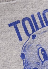 Szary materiał o miękkiej teksturze, z wyraźnym niebieskim napisem "DOTKNIJ" oraz niebieską twarzą postaci z kreskówki, częściowo zasłoniętą.