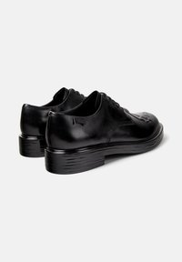 Un paio di scarpe eleganti in pelle nera con lacci, dotate di suole spesse e testurizzate e un logo sottile sul lato, adagiate su uno sfondo semplice.