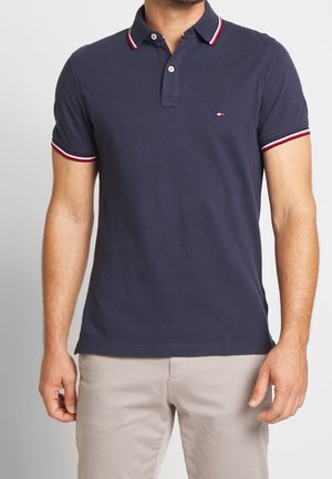 Mężczyzna ubrany w granatowe polo z krótkim rękawem z kołnierzykiem i wykończeniem rękawów w czerwono-białe paski, w połączeniu z jasnobeżowymi spodniami.