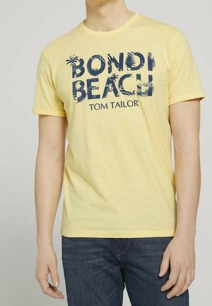 Homme portant un t-shirt jaune à manches courtes avec le texte "BONDI BEACH TOM TAILOR" et des graphiques de palmiers, accompagné d'un jean bleu foncé.