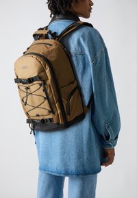 Bruine rugzak met zwarte accenten, voorzien van verstelbare schouderbanden, een externe bungeekoord en ritszakken, gedragen door iemand in denim.