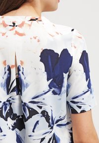 Blusa floreale in tessuto leggero, caratterizzata da motivi blu e bianchi con accenti arancioni, maniche corte e dettaglio con spacco sul retro.