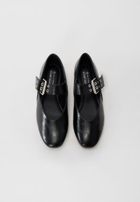 Schwarze vegane Lederschuhe mit einer abgerundeten Spitze, ausgestattet mit einem breiten Riemen mit einer metallischen Schnalle und Ösen. Glatte, leicht glänzende Textur.