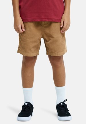 Dziecko ubrane w brązowe sztruksowe spodenki, białe skarpetki do kostek, czarne trampki i czerwoną koszulkę, z rękami swobodnie opuszczonymi wzdłuż ciała.