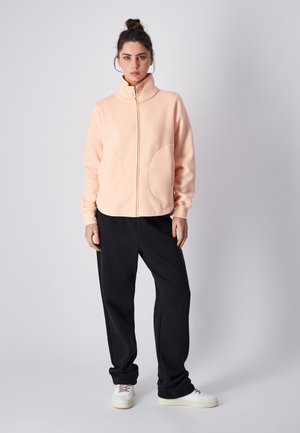 Sudadera con cremallera color melocotón y cuello alto, con bolsillos grandes y puños de canalé, combinada con pantalones negros holgados y zapatillas blancas.