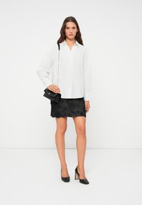 Camicia bianca con bottoni, mini gonna in pizzo floreale nero, scarpe con tacco nere e una piccola borsa nera con tracolla a catena.