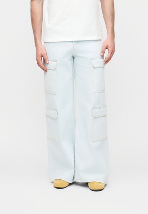 Person iført lyseblå vidbukser med store sidelommer og gule Puma-sneakers, stående mot en hvit bakgrunn.