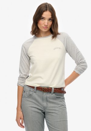Jonge vrouw met bruin haar, gekleed in een crèmekleurig en grijs lang mouw raglan shirt en gestreepte broek met een bruin gevlochten riem.