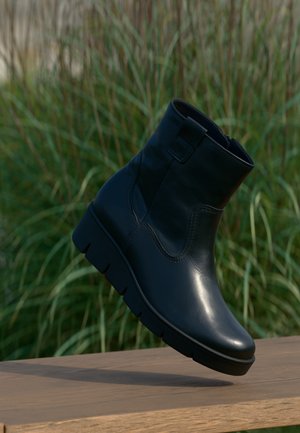 Bottine en cuir noir avec semelle épaisse texturée, exposée en extérieur sur une surface en bois avec un fond d'herbe verte.