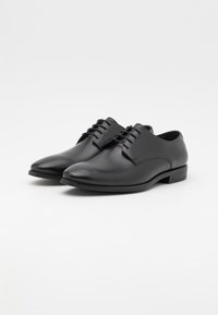 Chaussures habillées en cuir noir avec une texture lisse, bout arrondi et un laçage à six œillets. Semelle plate et détails minimalistes.