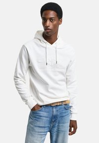 Sudadera blanca con cordón y logo en relieve, combinada con jeans de color azul claro. La tela parece suave y tiene un corte relajado.