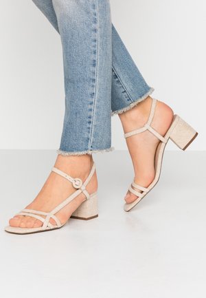 Fødder iført lyse beige remsandaler med blokhæl kombineret med lyseblå jeans med frynset kant på hvidt gulvbaggrund.