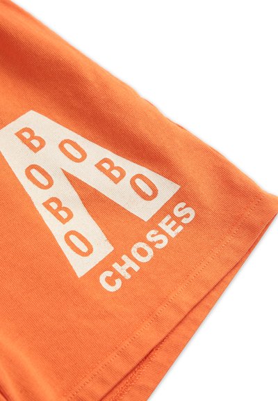 Oranžové bavlnené tričko s výrazným bielym textom "BOBO CHOSES" a štýlovým dizajnom písmena "A". Hladká textúra, krátke rukávy, okrúhly golier.