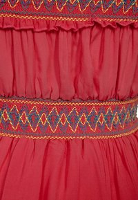 Tela roja con textura fruncida, presenta una cinturilla bordada colorida en patrones geométricos azules y amarillos. Bordes con volantes en la parte superior.