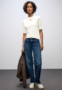 Top bianco a maniche corte con dettaglio a chiave, abbinato a jeans blu in denim, sneakers beige e una giacca marrone appoggiata su un braccio.