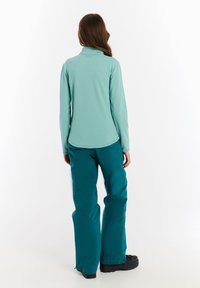 Svetlo mätová blúzka s dlhým rukávom a vysokým golierom, spárovaná s tmavo mätovými nohavicami s širokými nohavicami a čiernou obuvou. Hladká textúra, minimalistický dizajn.