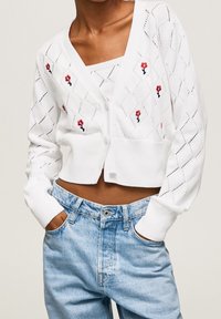 Bílý pletený cardigan s diamantovými dírkami, červenou květinovou výšivkou, krátký střih, nošený přes laděný bílý top a doplněný světle modrými džíny.