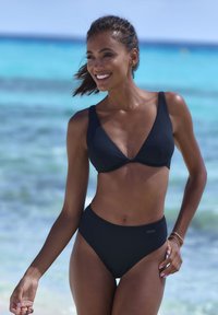 Bikini nero a coste con top triangolare e spalline larghe, accompagnato da uno slip a vita alta. Texture liscia su uno sfondo da spiaggia.