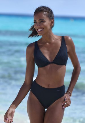 Melna ribota bikini, kuram ir trīsstūra formas krūšturis ar platām siksnām un augstiem vidukļa apakšbiksēm. Gluda tekstūra pludmales fonā.