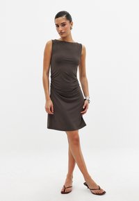 Robe sans manches marron avec des détails froncés sur le côté, longueur genou et tissu lisse. Le mannequin porte des tongs marron et un bracelet en argent.